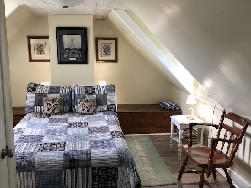 A cozy bedroom with slanted ceilings, a patchwork quilt on the bed, and a wooden chair next to a small side table, typical of Tilghman Island accommodations.
