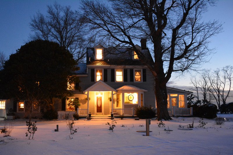 A two-story house at dusk with snow-covered ground and lights glowing in the windows, evocative of Black Walnut Point Inn discounts.
