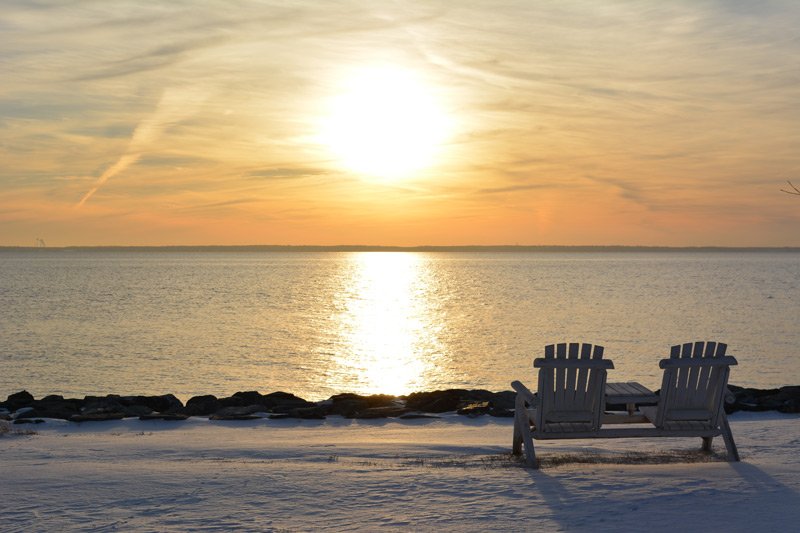 Two Adirondack chairs sit on a snow-covered shore at Black Walnut Point Inn, facing a sunset over the water.