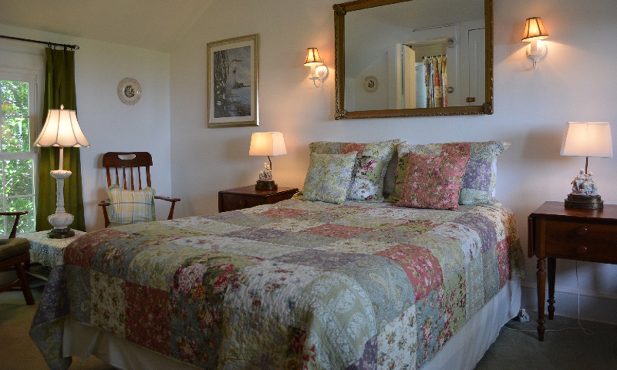A bedroom at a Tilghman Island Bed and Breakfast with a queen-sized bed featuring a floral patchwork quilt, two wooden nightstands with lamps, a wooden chair, and a large mirror hanging above the bed.