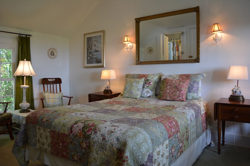 A bedroom featuring a patchwork quilt bed, wooden nightstands with lamps, a framed mirror, and a wooden chair by a window with green curtains, perfect for Tilghman Island accommodations.