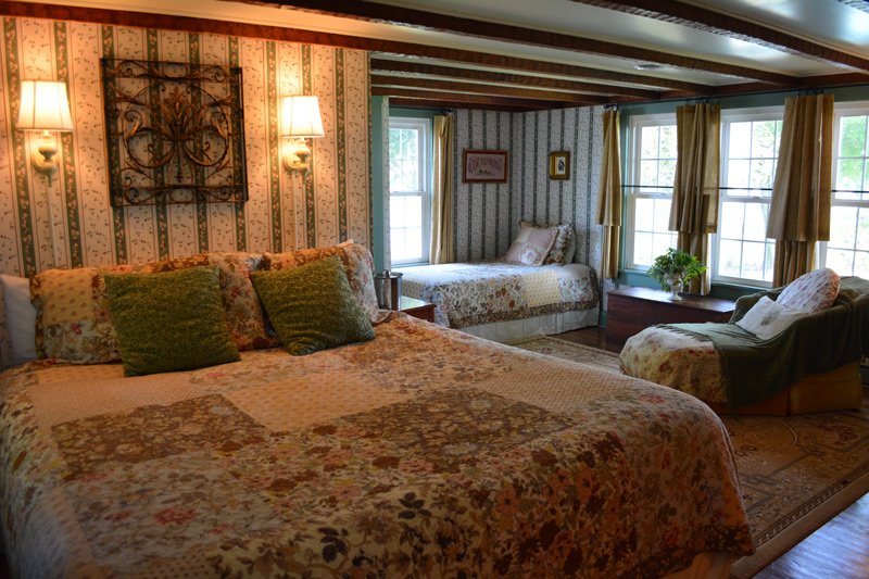 A bedroom with a large floral patchwork bed in the foreground, a single bed against the back wall, and an armchair near the windows, typical of Tilghman Island accommodations.