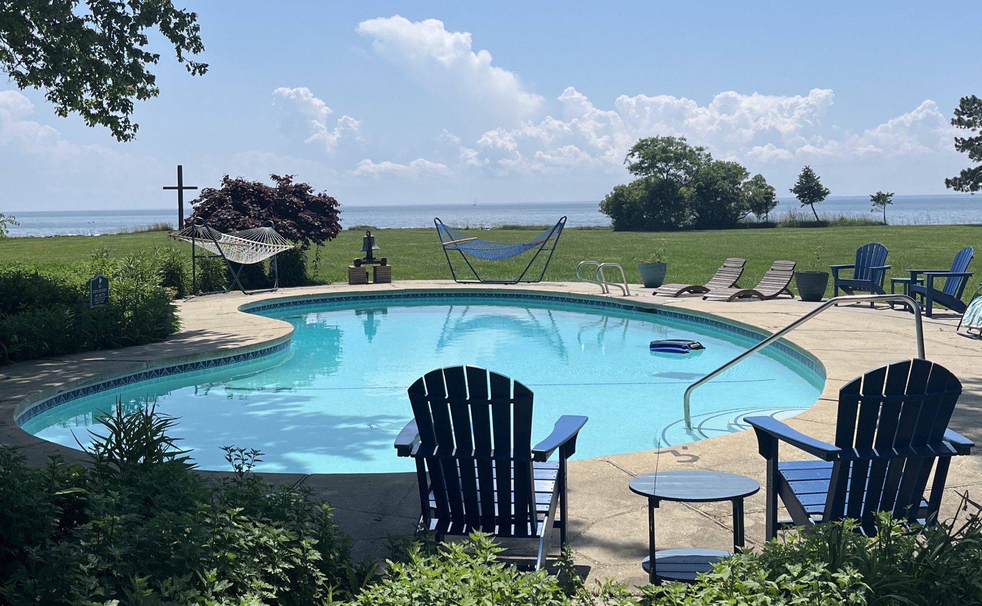 A swimming pool surrounded by blue Adirondack chairs and hammocks at a Tilghman Island bed and breakfast, overlooking a grassy lawn and the ocean under a blue sky.